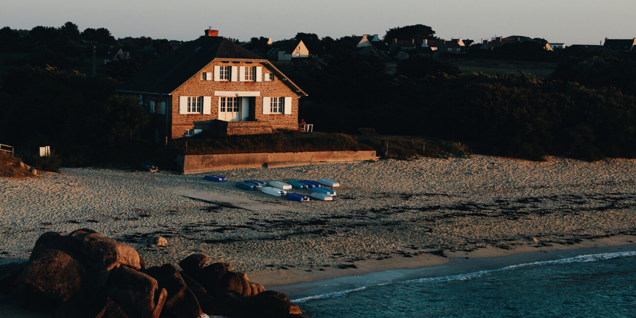 wide-shot-brown-house-sandy-seashore-by-sea-surrounded-with-rocks-trees California coast house representing second home and investment property differences