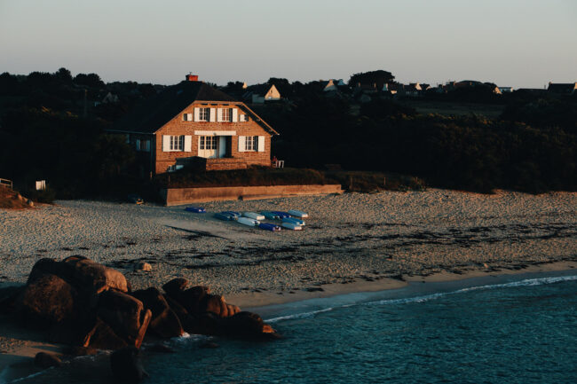 wide-shot-brown-house-sandy-seashore-by-sea-surrounded-with-rocks-trees California coast house representing second home and investment property differences